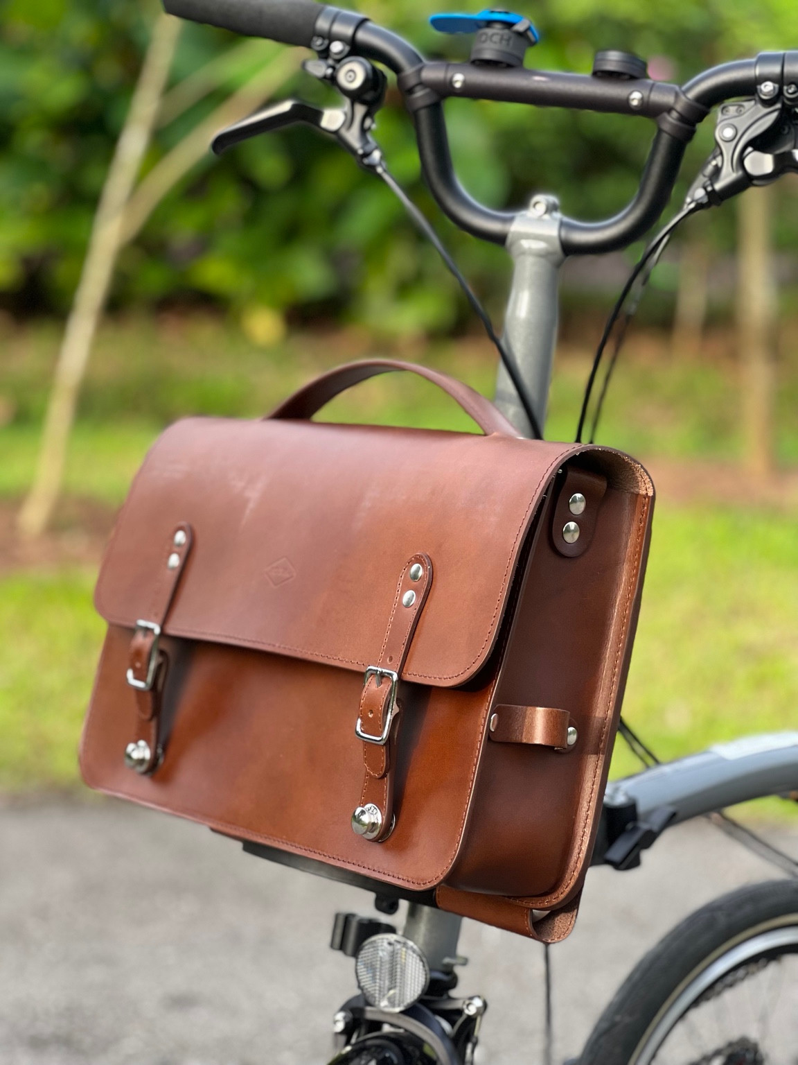 Three-quarter view of the brown Souma leather briefcase showing silver hardware and side strap holder