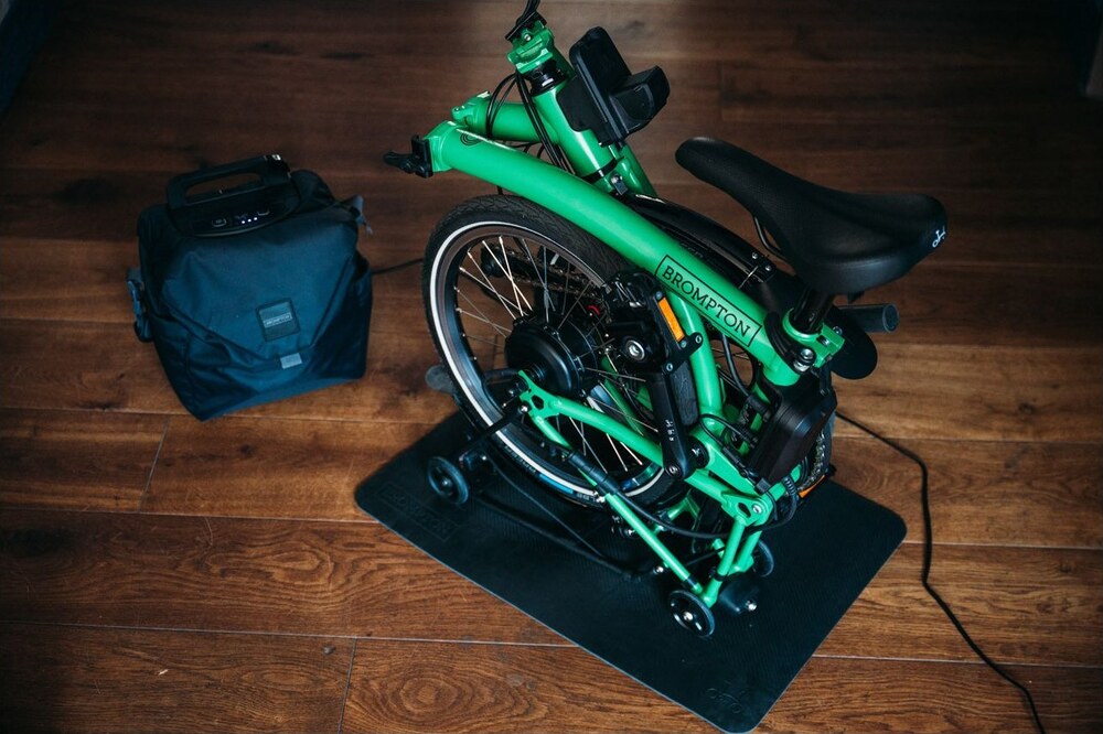 Folded green Brompton bike standing on a black Brompton Storage Mat indoors