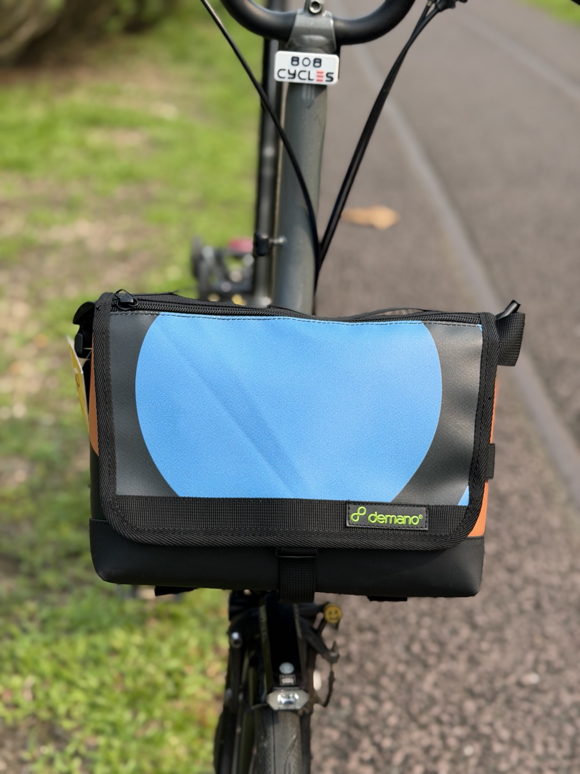 Front view of the actual Demano Ribera 1705 bag mounted on a Brompton, showing a bright blue circular graphic on charcoal and black trim.