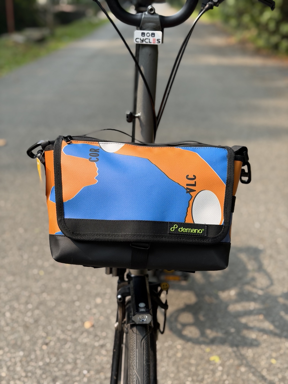 Front view of the actual Demano Ribera 1706 bag mounted on a Brompton, with blue and orange graphics, white accents, black lettering and black trim.