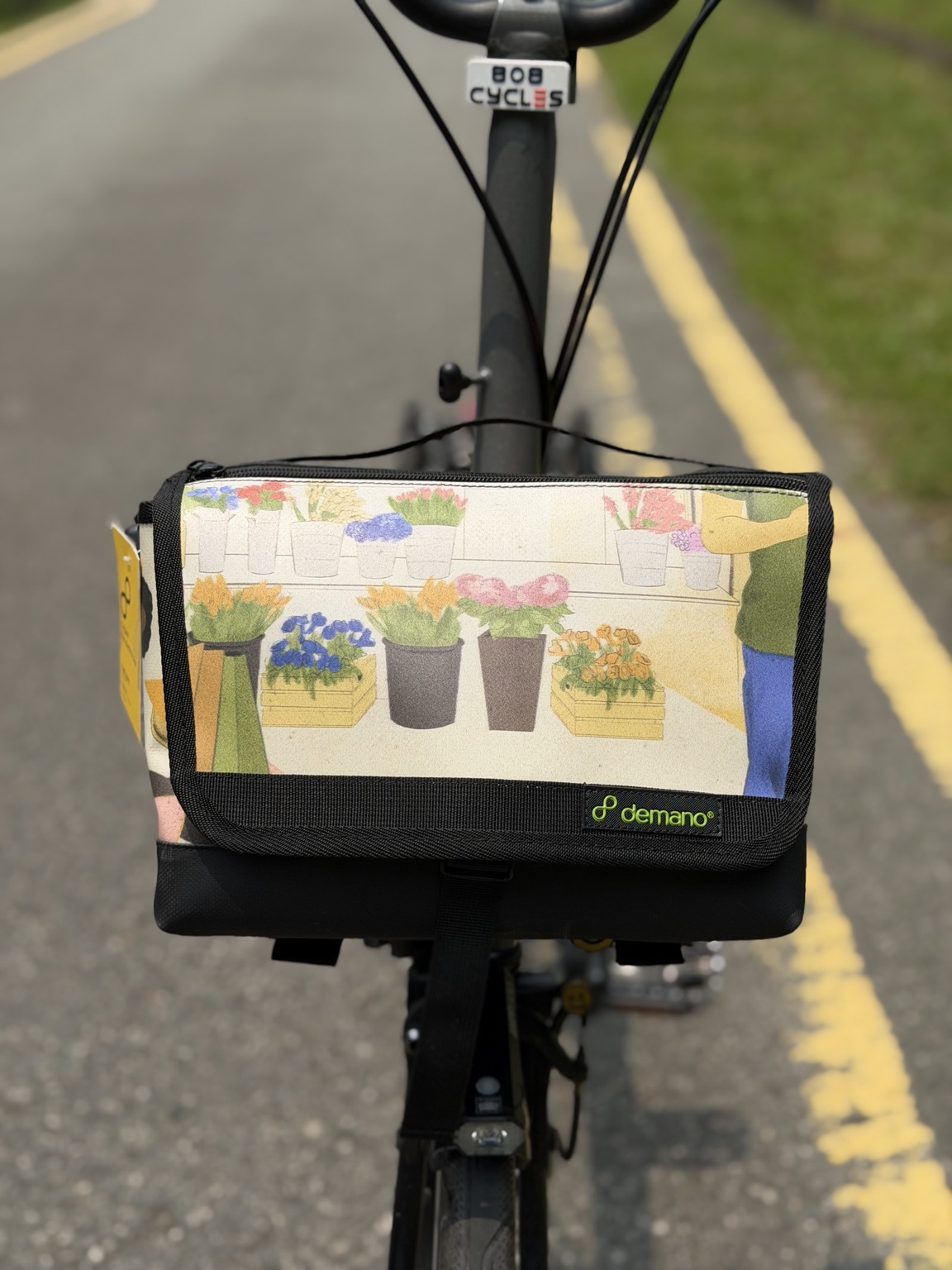 Front view of the actual Demano Ribera 1708 bag mounted on a Brompton, showing illustrated flower pots and crates on cream canvas with black trim.