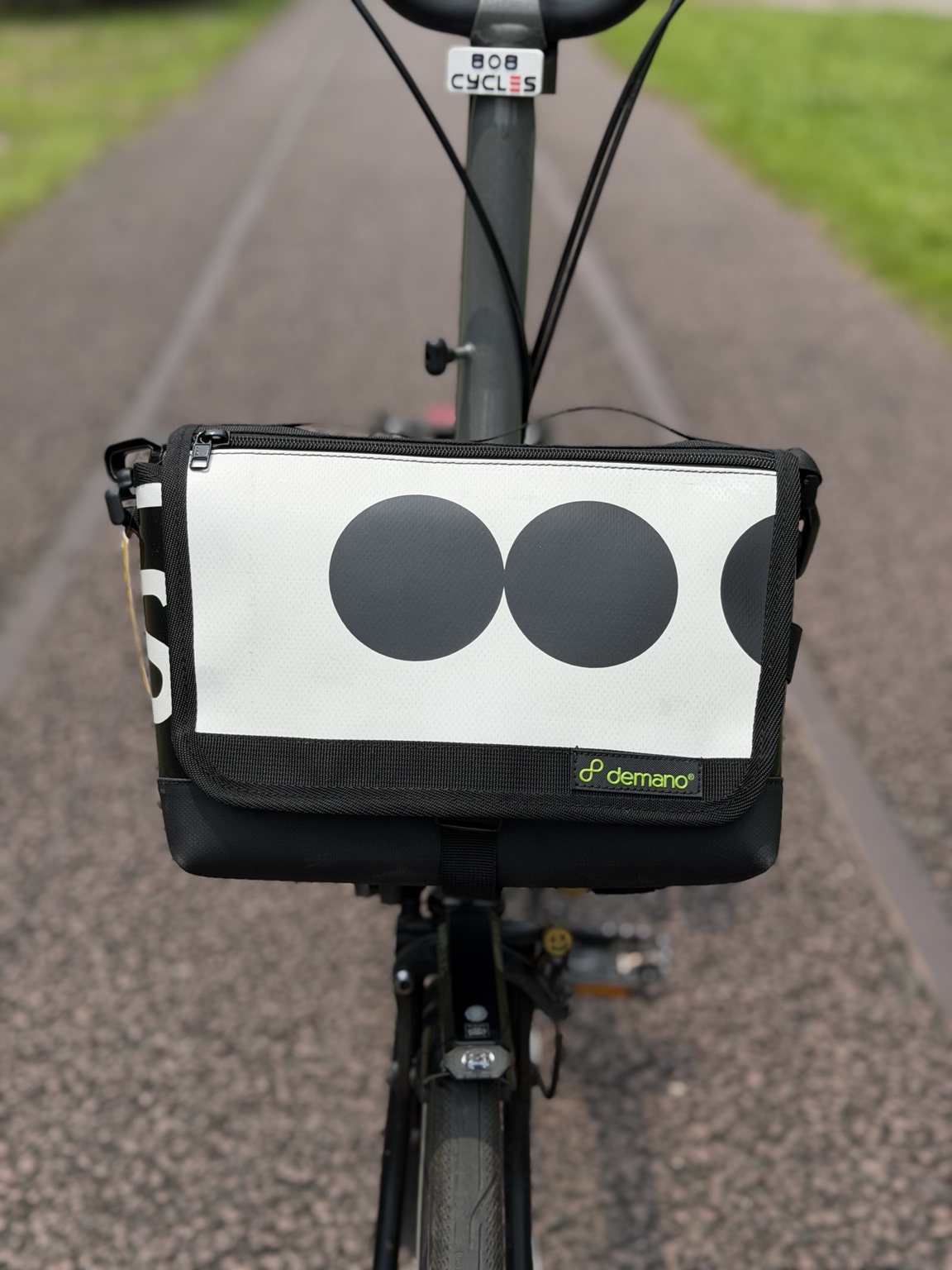 Front view of the actual Demano Ribera 1709 bag on a Brompton, showing two complete charcoal circles and a partial circle on the white flap with black trim.