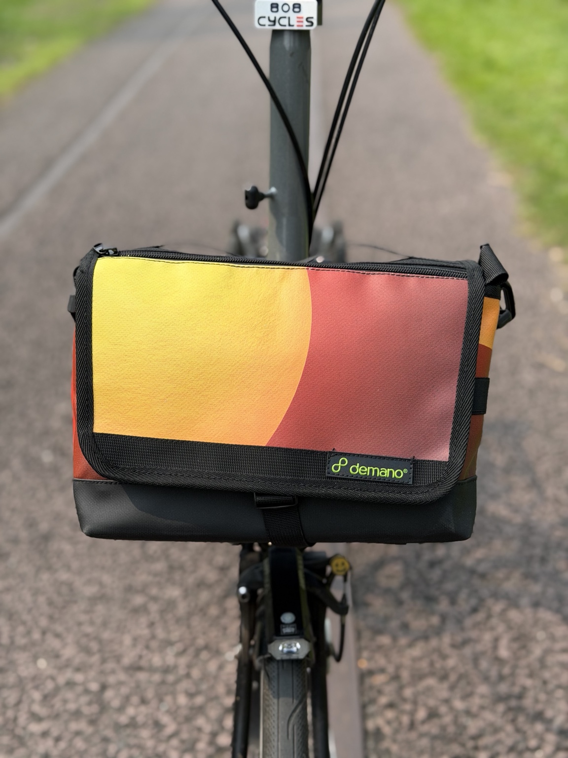 Front view of the actual Demano Ribera 1710 on a Brompton, showing the large curved yellow-orange block on its red-burgundy flap with black trim.