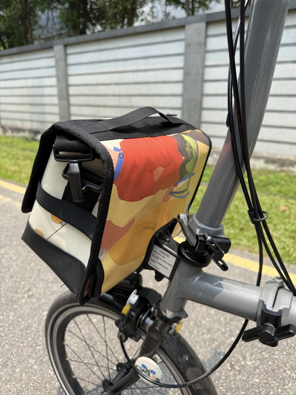 Rear-side angle of Demano Ribera 1708 showing a red-and-yellow illustrated figure and blue detail on the banner print beside the original Brompton stem and cables.